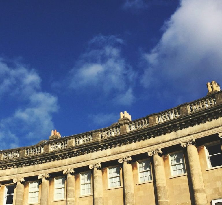 Photograph of the Royal Crescent in Bath, shown against a bright blue sky. The curved row of Georgian buildings is framed within a circular image.